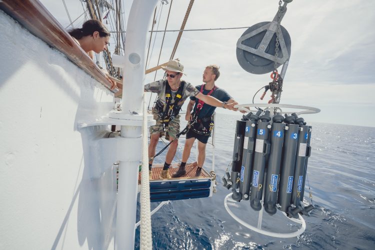 Two researchers stand on a platform off the side of the Statsraad Lehmkuhl, holding onto a piece of equipment that will be lowered into the water for water sampling. 