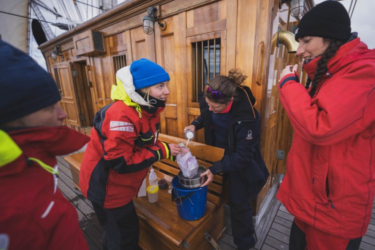 Four researchers stand on the top deck of the ship in waterproof jackets, working with a bucket and droppers. 