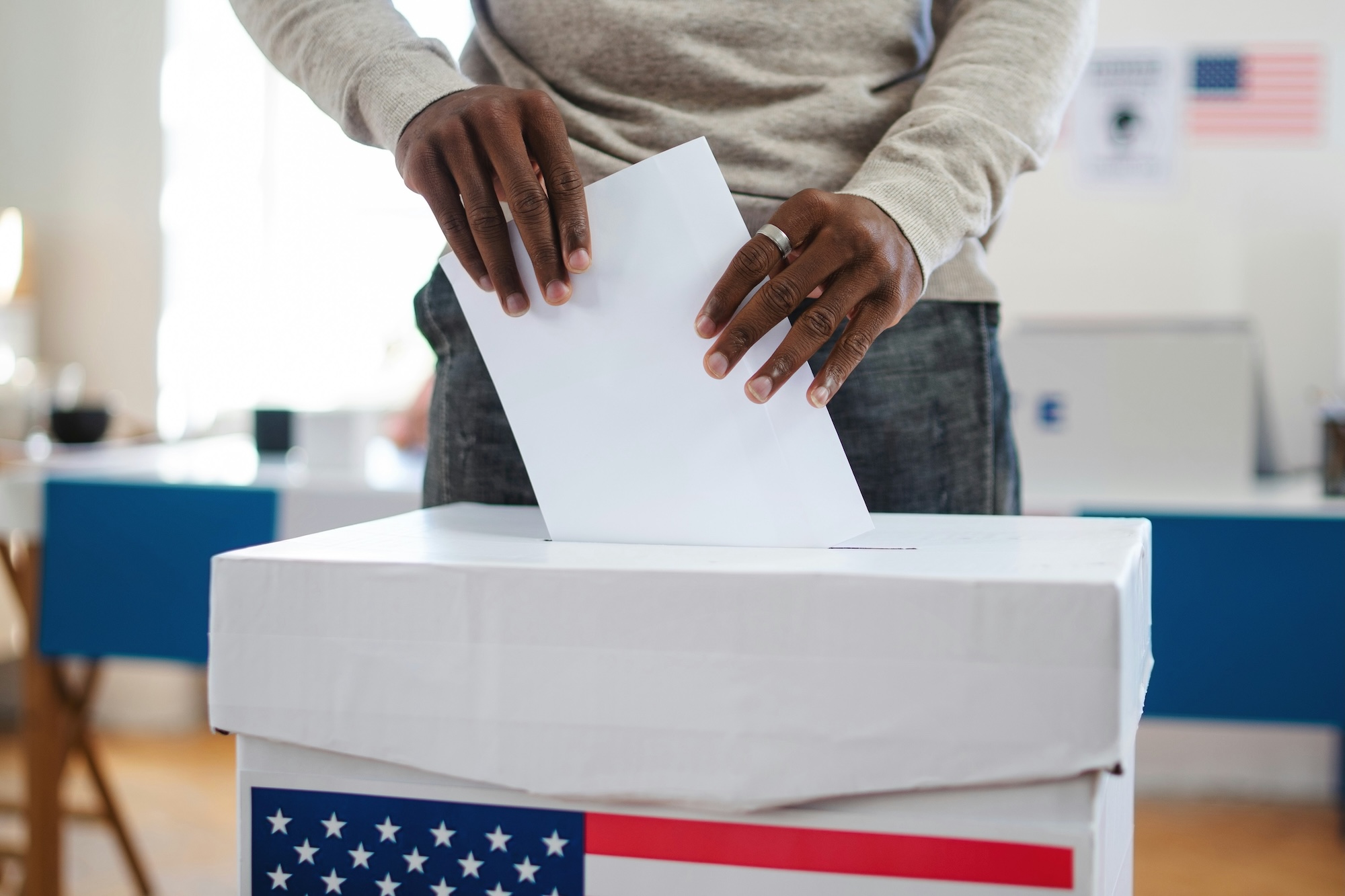 A Black person puts a ballot into a voting box with the United States flag on it.