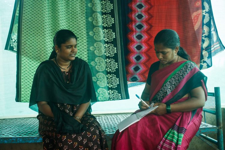 An Accredited Social Health Activist converses with a woman sitting next to her while filing out paperwork on a clipboard.