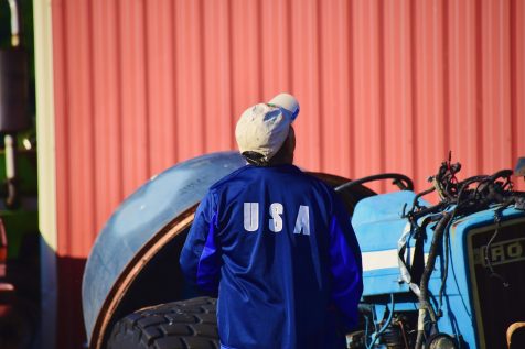 man working on a farm with USA printed on the back of his shirt and equipment and a red barn in the background