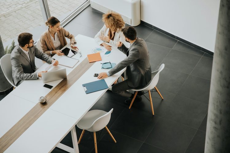 Four people sit around a board room table talking with scattered papers and electronics on the table in front of them.